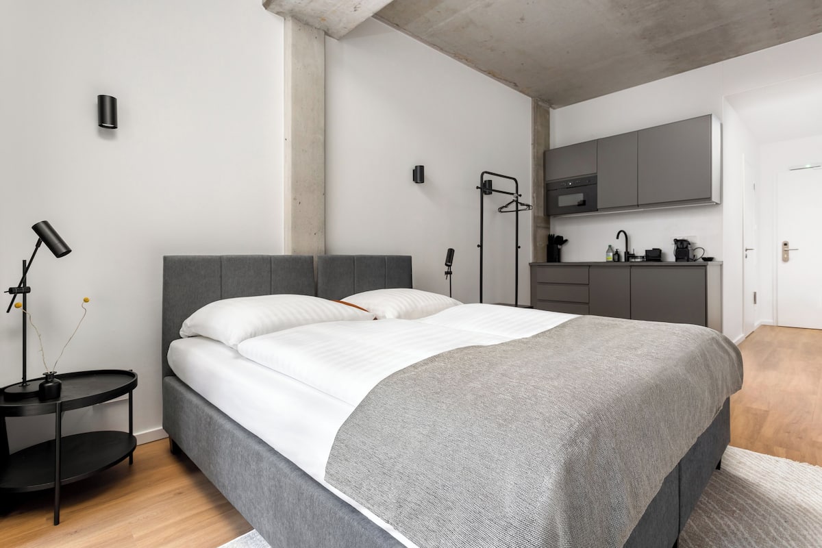 A modern bedroom with a grey bed, a bedside table with a lamp, and a kitchen area with cabinets and a sink.