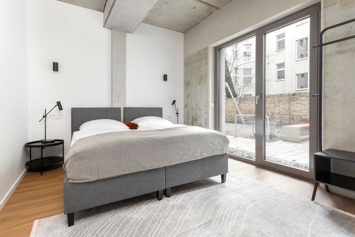 A modern bedroom with a grey bed, a large window, and a view of the outside.