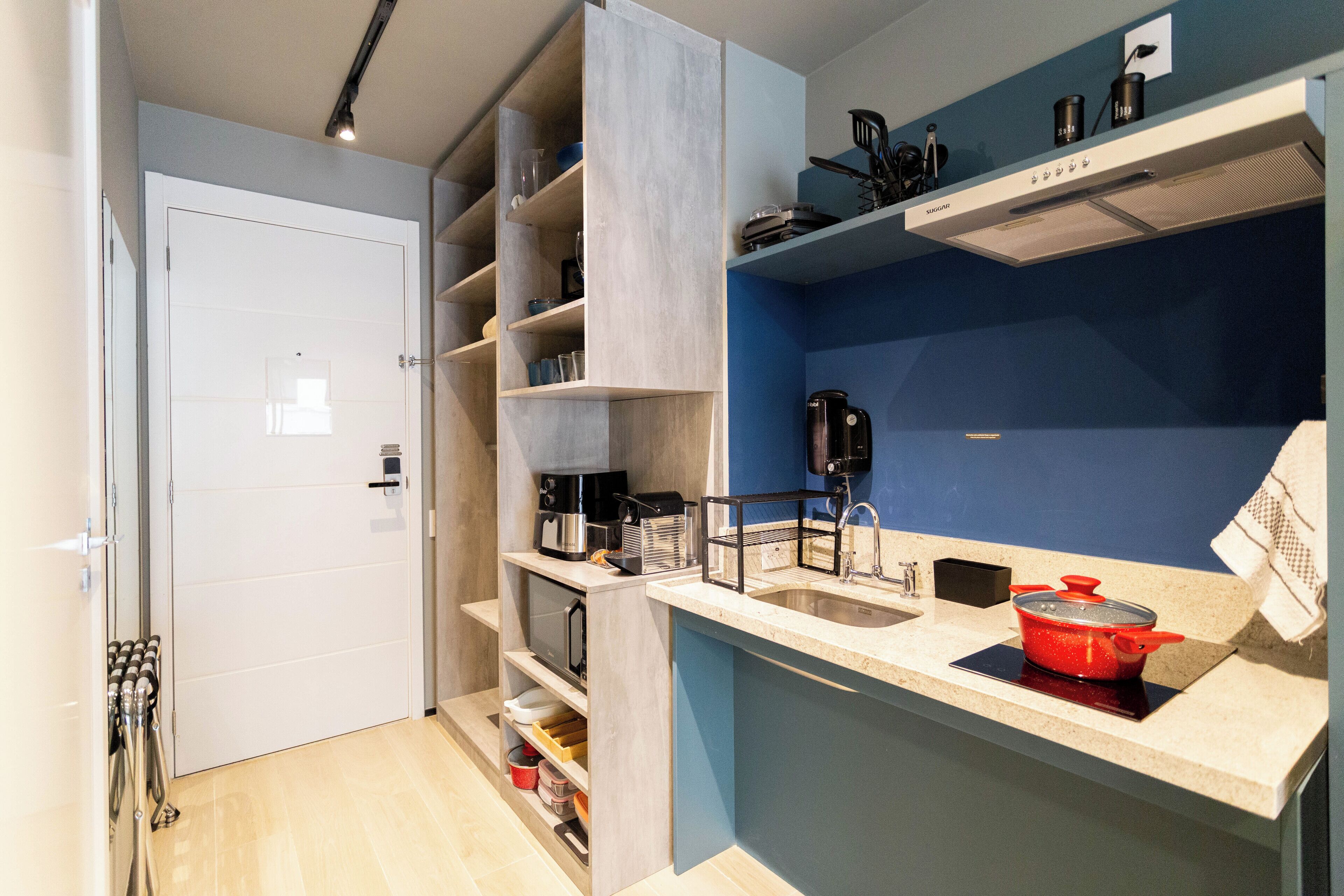 A compact kitchen with a built-in oven, a sink, and a microwave. There are shelves with various kitchen items and a red pot on the counter.