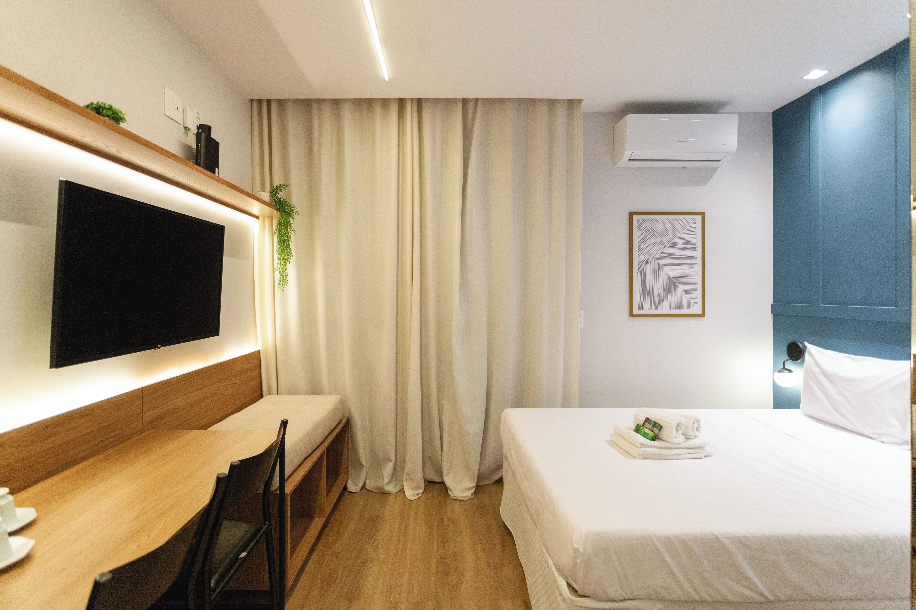 A hotel room with a flat-screen TV, a desk, a bed, and a window with curtains.