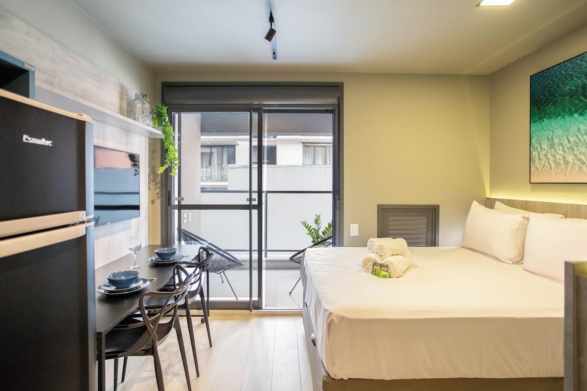 Een compacte hotelkamer met een bed, een kleine eettafel met stoelen, een koelkast en een schuifdeur naar het balkon.