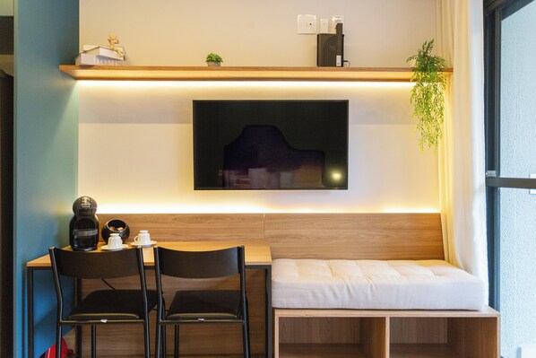 Een moderne kamer met een houten bureau, twee zwarte stoelen, een klein bankje, een hangend schilderij, een plank met een plant en een raam.