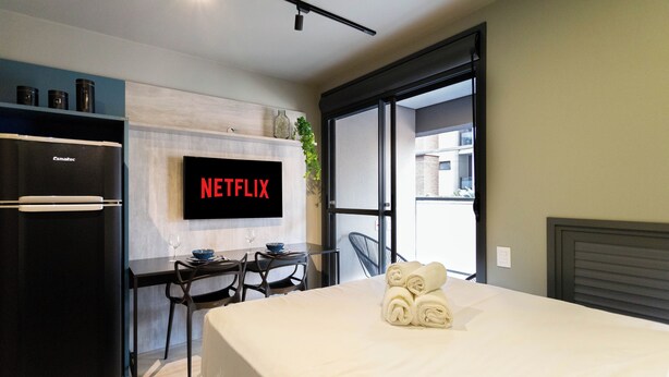 Una habitación de hotel moderna con una cama, un escritorio con una silla, un refrigerador negro, un televisor de pantalla plana mostrando "NETFLIX" y una ventana que ofrece vistas a edificios.