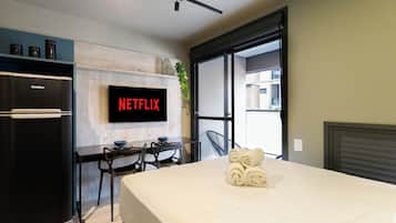 Una habitación de hotel moderna con una cama, un escritorio con una silla, un refrigerador negro, un televisor de pantalla plana mostrando "NETFLIX" y una ventana que ofrece vistas a edificios.
