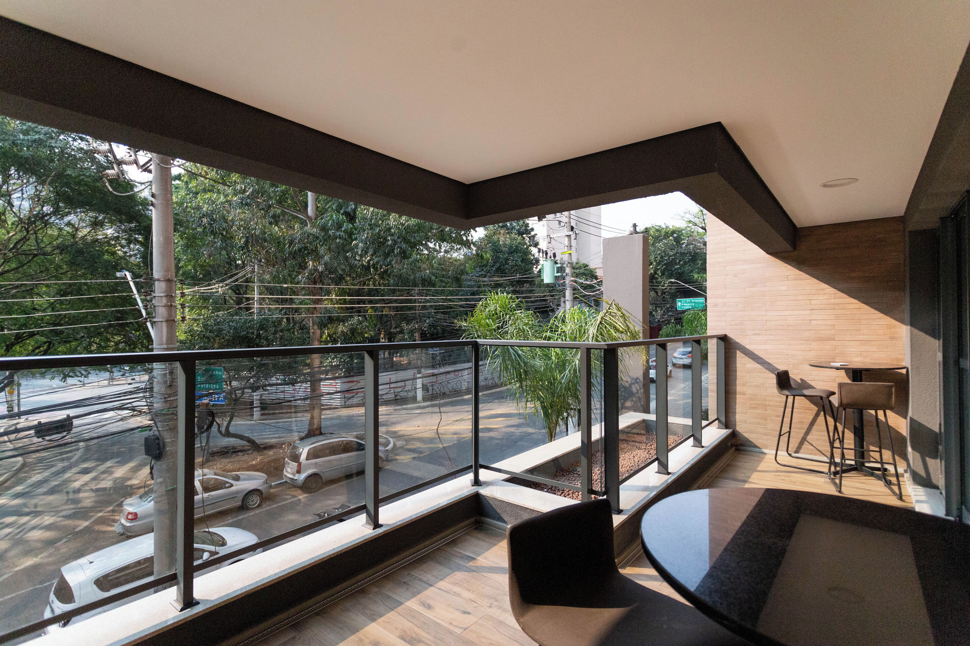 Un balcón moderno con barandilla de vidrio, una zona de estar con mesa y sillas, y vistas a la calle y al verdor.