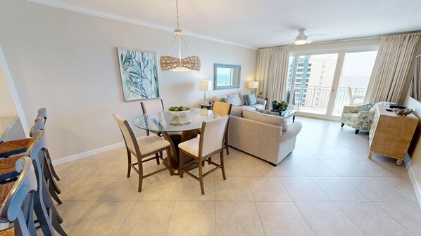 A modern living room with a glass dining table, two sofas, a coffee table, and a view of the city through large windows. - Sterling Shores 902 3 Bedroom Condo (Destin)