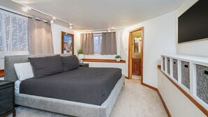Ett sovrum med en stor säng, ett sängbord, en liten avlastningsyta, ett fönster med gardiner, en väggmonterad TV och en dörr som leder till ett annat rum.
