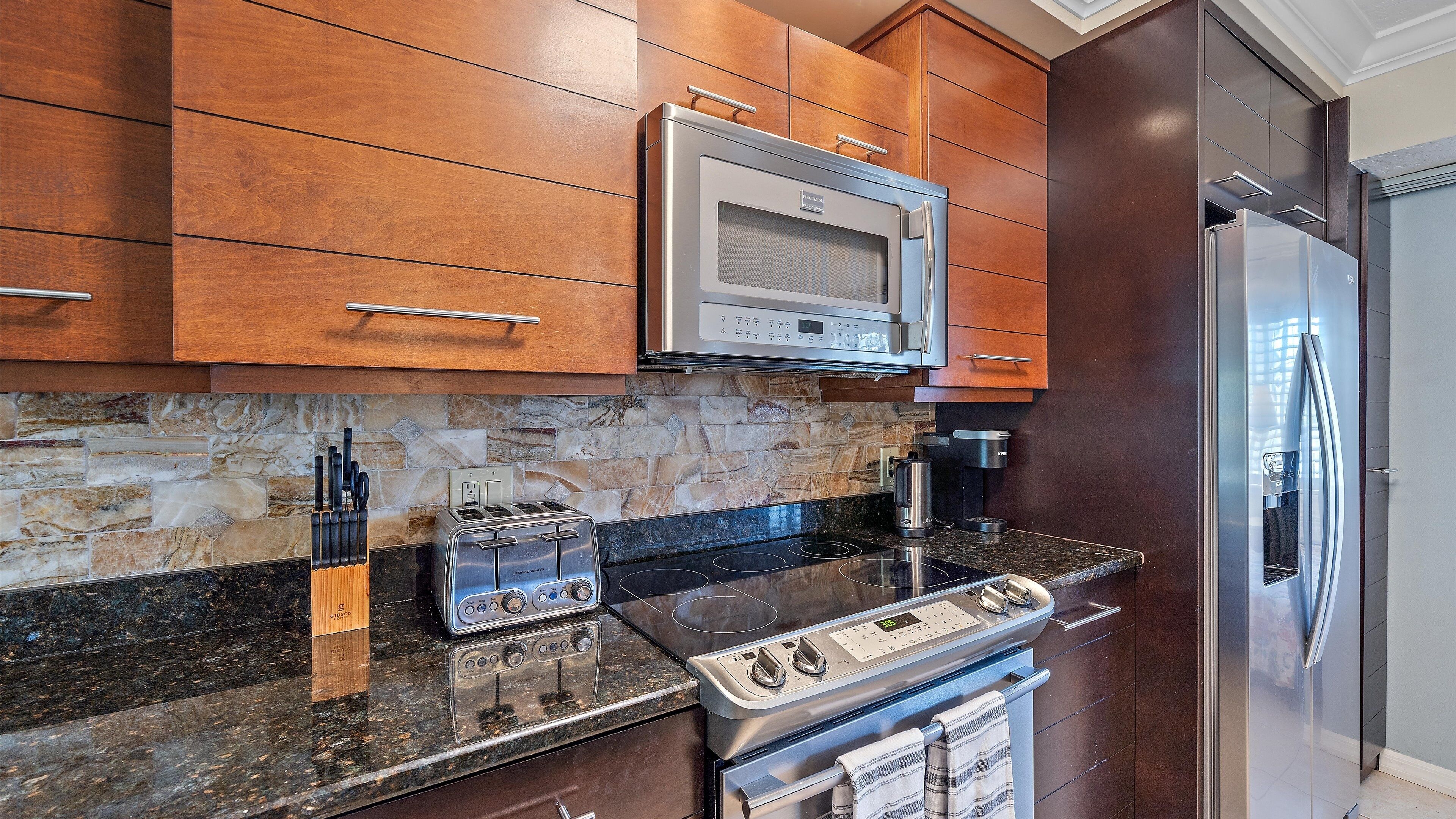 A modern kitchen with wooden cabinets, a stone backsplash, a stainless steel range, a built-in microwave, and a refrigerator.