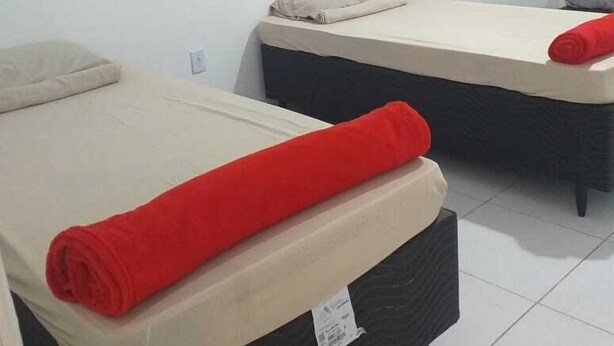 Dos camas individuales con toallas rojas, una obra de arte pequeña en la pared y un cojín gris sobre un suelo de baldosas blancas.