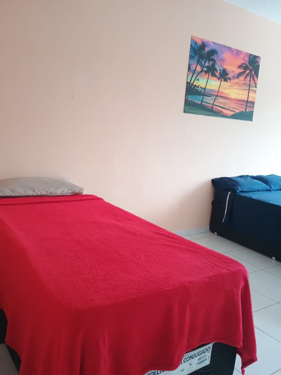 A room with a bed covered in a red blanket, a couch with blue cushions, and a wall-mounted painting of palm trees and a sunset.