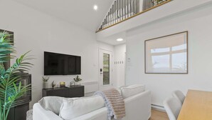 A modern living room with a white sofa, a wooden dining table, and a staircase leading to an upper level. - Spa | Mountain | Modern Comfort | Elska (Saint-Côme)