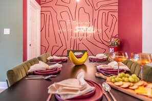 Un comedor con una mesa larga dispuesta para seis personas, con un diseño moderno que incluye un patrón geométrico en la pared, un cuenco decorativo y una bandeja de frutas.