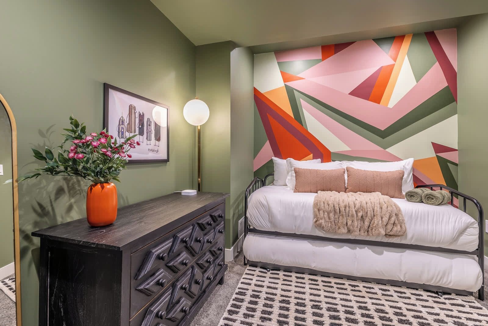 A bedroom with a bed, a dresser, a vase with flowers, a framed picture, and a geometric wall design.