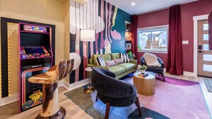 Un salon moderne avec un canapé vert, un fauteuil noir et une sculpture en bois. On aperçoit une borne d’arcade colorée sur la gauche et une fenêtre donnant sur des bâtiments à l’extérieur.