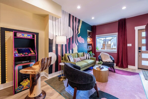 Una sala de estar moderna con un sofá verde, un sillón negro y una escultura de madera. A la izquierda, una máquina recreativa colorida y una ventana con vistas a edificios.