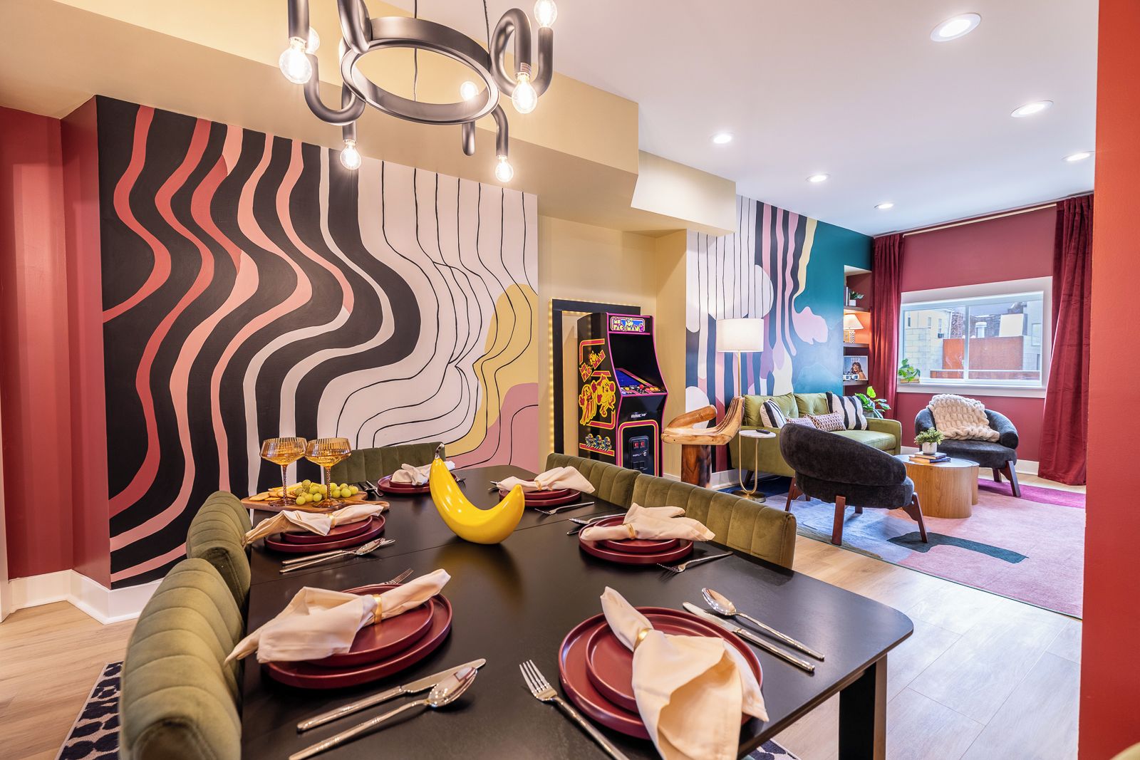 A dining area with a long table set for a meal, a colorful abstract wall, and a retro arcade machine in the background.