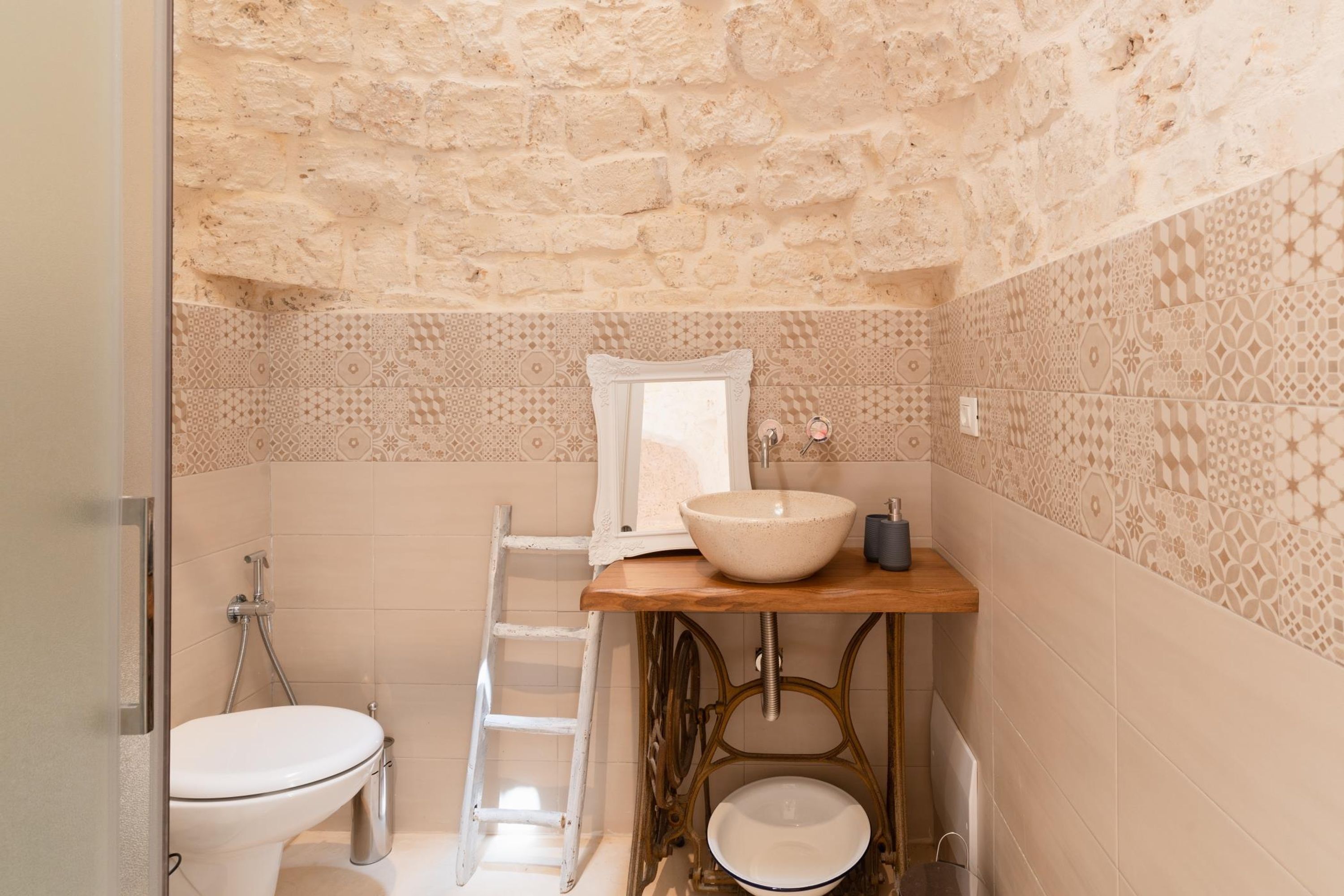 A bathroom with a stone wall, a wooden vanity, a white sink, a mirror, a toilet, and a shower.