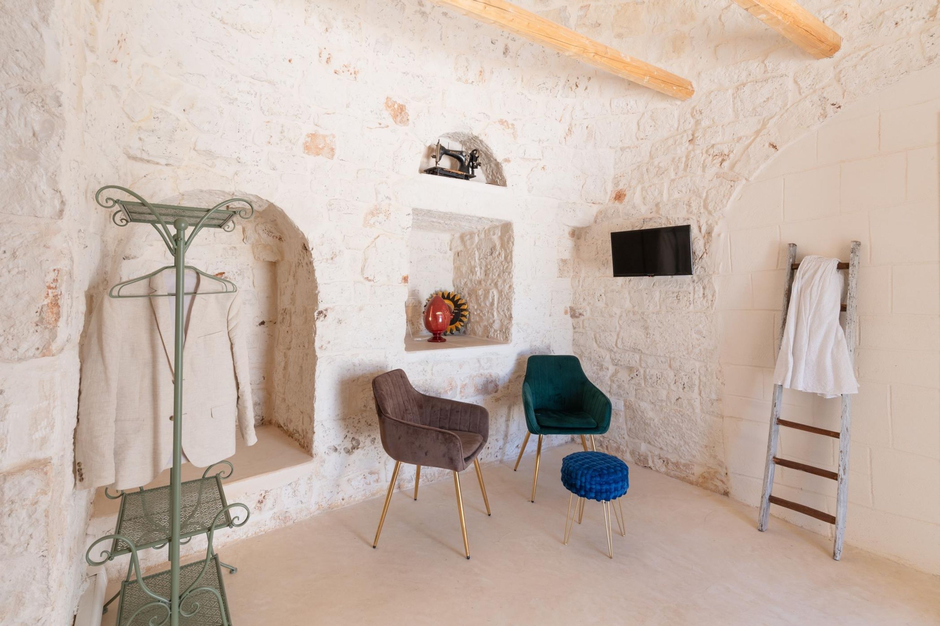 A rustic room with a stone wall, a metal coat rack, three chairs, a small shelf with a decorative item, a ladder, and a flat-screen TV mounted on the wall.