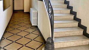 Un escalier avec une rampe noire, menant à l’étage supérieur. Le sol est recouvert de carreaux formant un motif géométrique.
