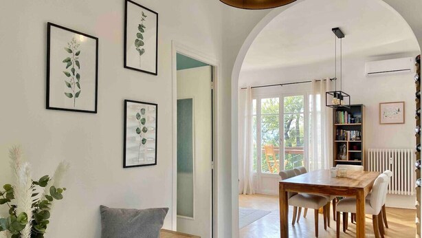 Un salon moderne avec une table à manger en bois, un canapé avec un coussin gris et des reproductions botaniques encadrées accrochées au mur.