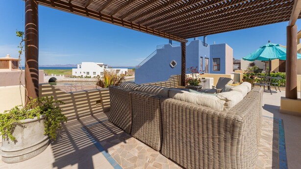 A covered outdoor seating area with wicker furniture, a view of the sea, and buildings in the background.
