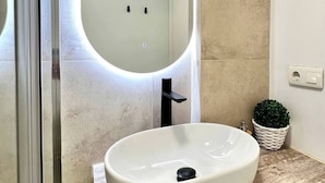 A bathroom with a large oval sink, a mirror with integrated lighting, a small potted plant, and a visible electrical outlet. - Modern apartment in Juan de Austria (Málaga)