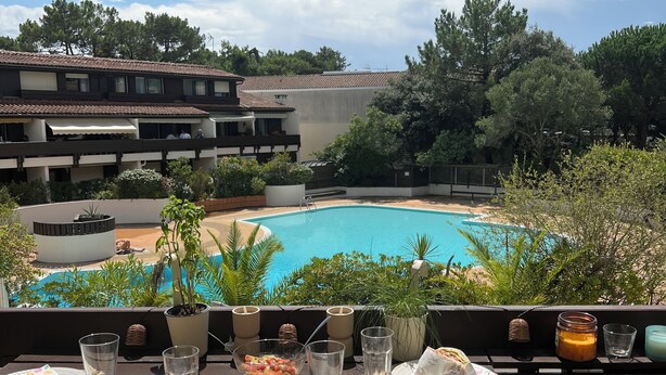 An outdoor dining area with a view of a swimming pool, surrounded by greenery and a building with multiple balconies.