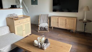 A living room with a sofa, a coffee table with candles and pinecones, a wicker chair, a wicker cabinet, a TV on the wall, and a lamp.
