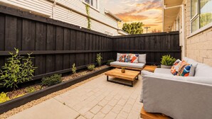 Un patio trasero con cerco de madera, un sofá gris, una mesa de madera y un pequeño jardín.