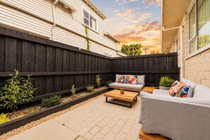 Un patio trasero con cerco de madera, un sofá gris, una mesa de madera y un pequeño jardín.
