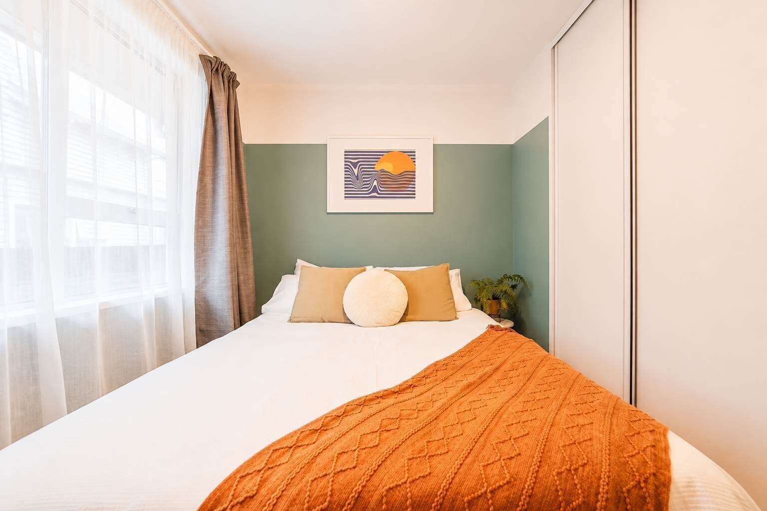 A hotel room with a bed, a window with curtains, a wall-mounted artwork, and a bedside plant.