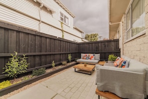 A backyard with a wooden fence, a gray sofa, and a wooden coffee table. - Hereford Haven 2BR w Parking | Near CBD & Stadium (Christchurch)