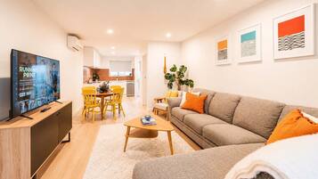 A modern living room with a flat-screen TV, a sofa, a coffee table, and a dining area with chairs.