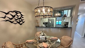 A dining area with a round glass table, four wicker chairs, and a central chandelier. - Bayfront Condo: Your Sunset Paradise on Fort Myers Beach (Fort Myers Beach)