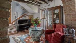 A living room with a piano, a red patterned armchair, a glass table with a vase, and a cabinet with glass doors.