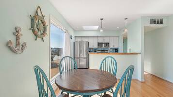 A dining area with a round wooden table and turquoise chairs, a kitchen with stainless steel appliances, and a view through a glass door.