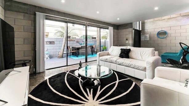 A modern living room with a white sofa, a glass coffee table, and a view of an outdoor pool area. - Las Olas Triplex w/ Water View, 5min to Beach (Fort Lauderdale)