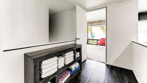 A modern hotel room with a dark wood console table holding neatly folded towels and a striped blanket. A white door leads to an outdoor area with a red chair and a view of greenery. - Las Olas Triplex w/ Water View, 5min to Beach (Fort Lauderdale)