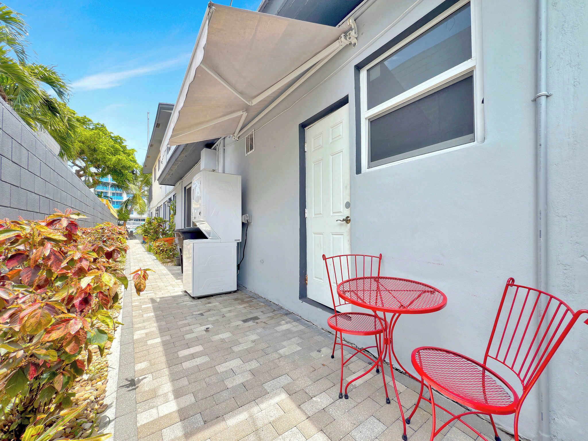 Une petite terrasse extérieure, dotée d’un auvent blanc, d’un réfrigérateur blanc et de chaises en métal rouge.