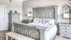 Un dormitorio con una cama grande, dos mesitas de noche, un banco, un aparador y dos lámparas de pared.