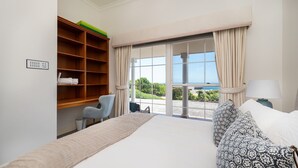 Une chambre d’hôtel avec un grand lit, une étagère en bois où sont posées des serviettes, une chaise, une lampe et une vue sur la mer par une porte vitrée.