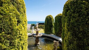 Un jardin magnifiquement entretenu, avec une fontaine centrale, bordé de haies soigneusement taillées, et offrant une vue sur la mer en arrière-plan.