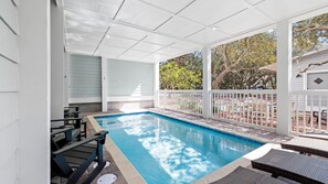 A covered outdoor swimming pool with a white fence, lounge chairs, and a view of trees and a house in the background.