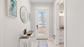 A hallway with a white door, a mirror, a welcome sign, a potted plant, and a view of the outdoors.
