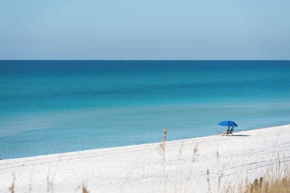 A beach with a clear blue sky and calm sea. There is a person sitting under a blue umbrella on the sand.