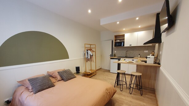 Une chambre compacte comprenant un lit, une table de chevet et un téléviseur fixé au mur. À côté se trouve une petite cuisine équipée d’un four à micro-ondes, d’un évier et de quelques tabourets de bar.
