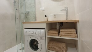 Une petite salle de bain dotée d’une douche à parois de verre, d’un lave-linge, d’un lavabo avec un miroir au-dessus et d’étagères où sont rangées les serviettes.