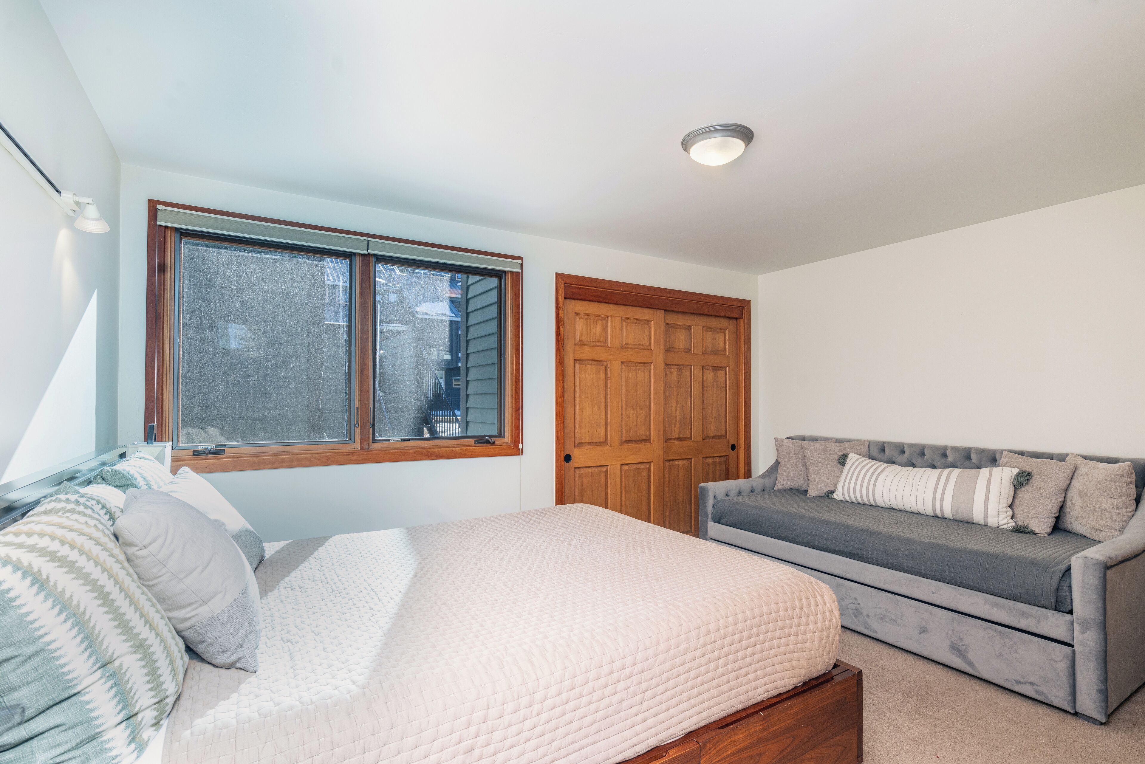 A bedroom with a bed, a grey sofa, a wooden wardrobe, and a window.