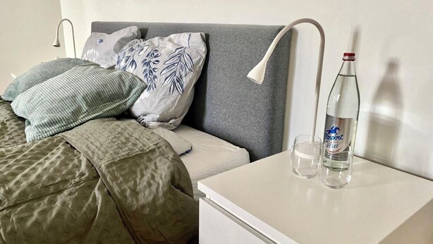 A neatly made bed with a grey headboard, a green patterned blanket, and several pillows, including one with a floral design. A white bedside table holds a bottle and a glass. - Cozy room in Remich (Remich)