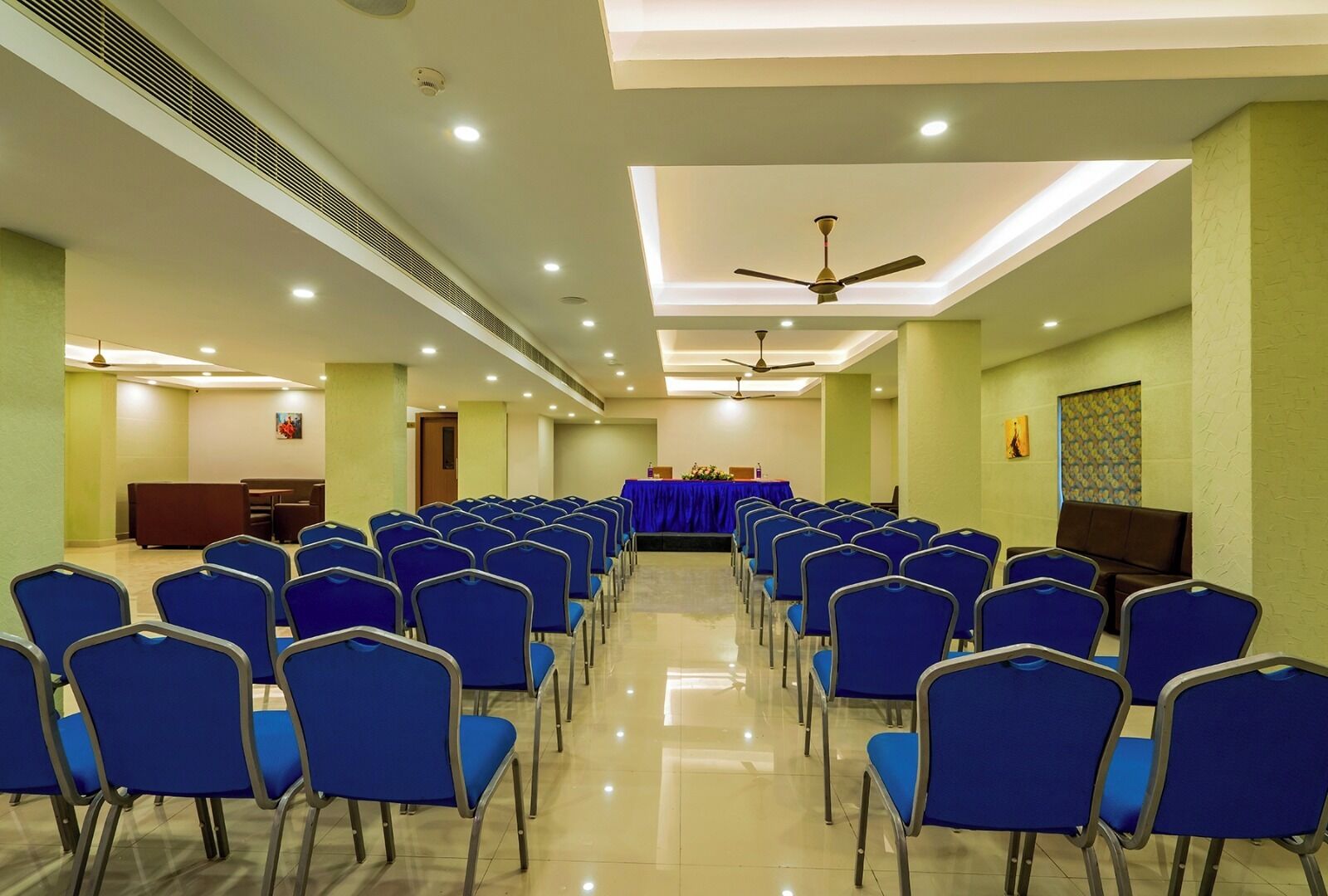 A conference room with rows of blue chairs, a stage with a blue tablecloth, ceiling fans, and recessed lighting.
